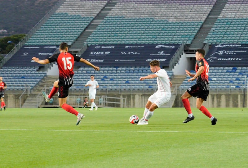 A football player in a white kit kicking a ball with a player in a red and black kit attempting to block the shot