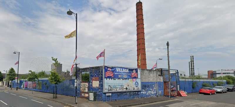 Street view of vacant site surrounded by blue hoarding with a tall red brick chimney in the background.