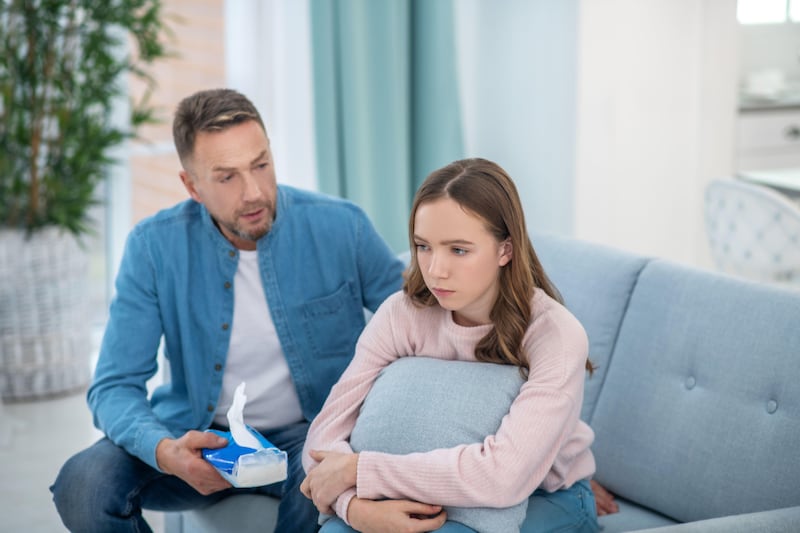 Concerned dad sitting on a sofa with his daughter offering her a box of tissues