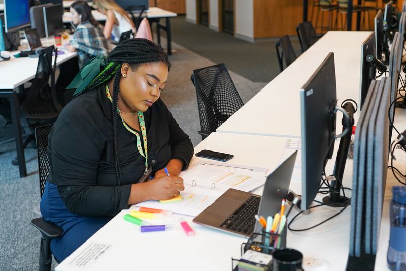 Person wearing sunflower lanyard to show less visible disabilities sits at desk writing on post-it note, with coloured highlighters and a notepad on the desk and an open plan office behind them. (Business Disability Forum)