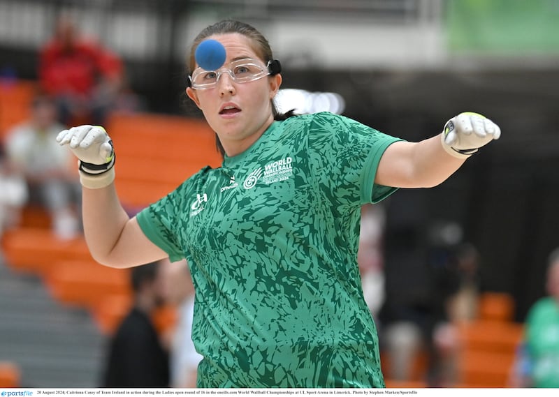 A woman wearing a green top, white gloves and clear glasses looking at a blue top winding up to strike the ball with her hand