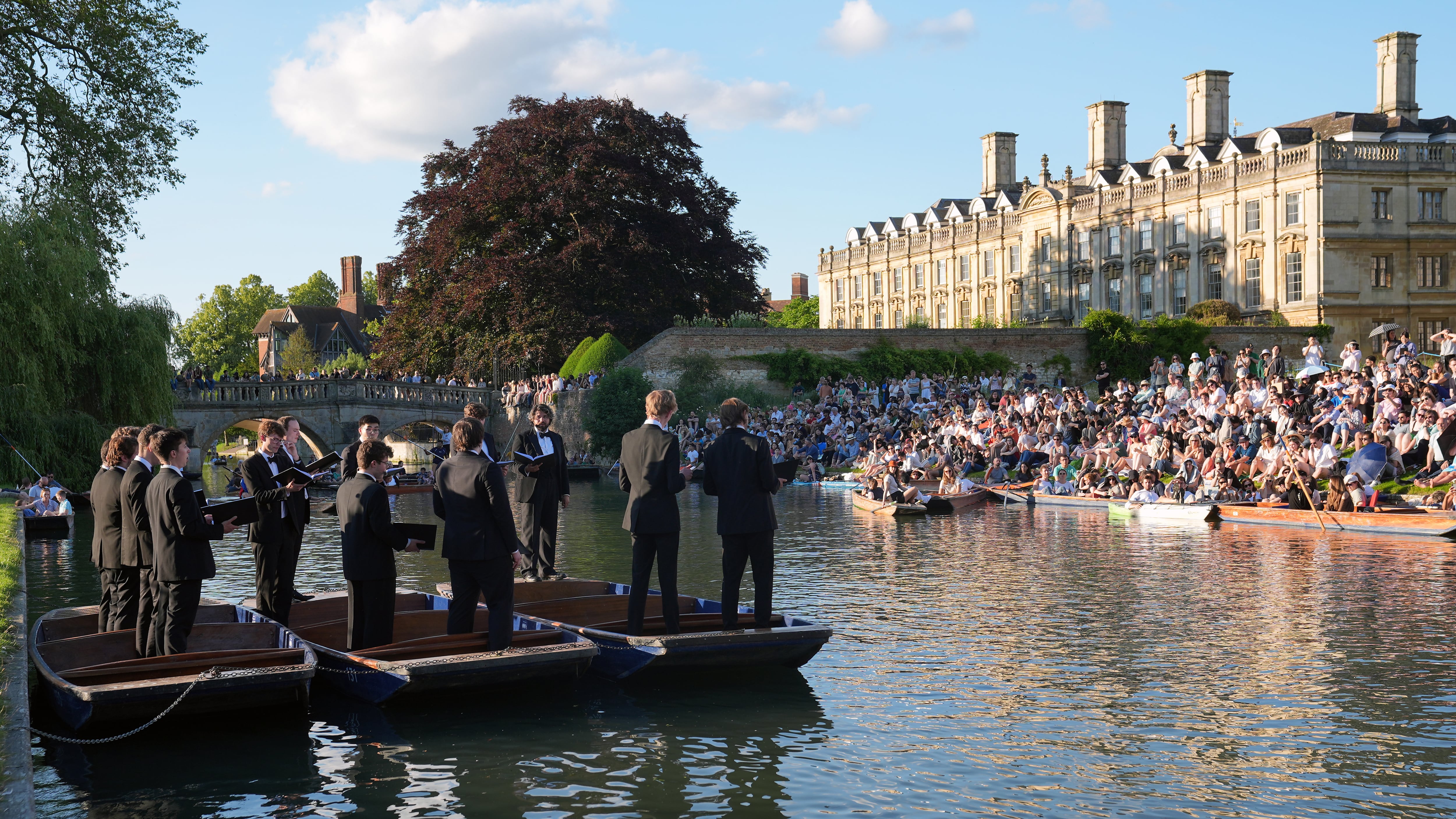 People sit on the banks of the River Cam and lawns of King’s College in Cambridge, as they listen to The King’s Men perform