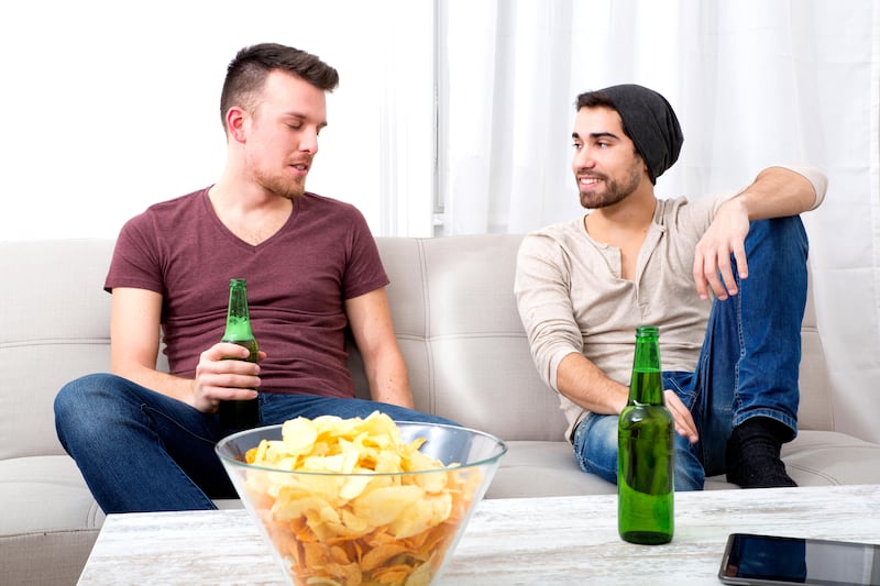 Two male friends drinking beer and eating crisps having a conversation in a living room