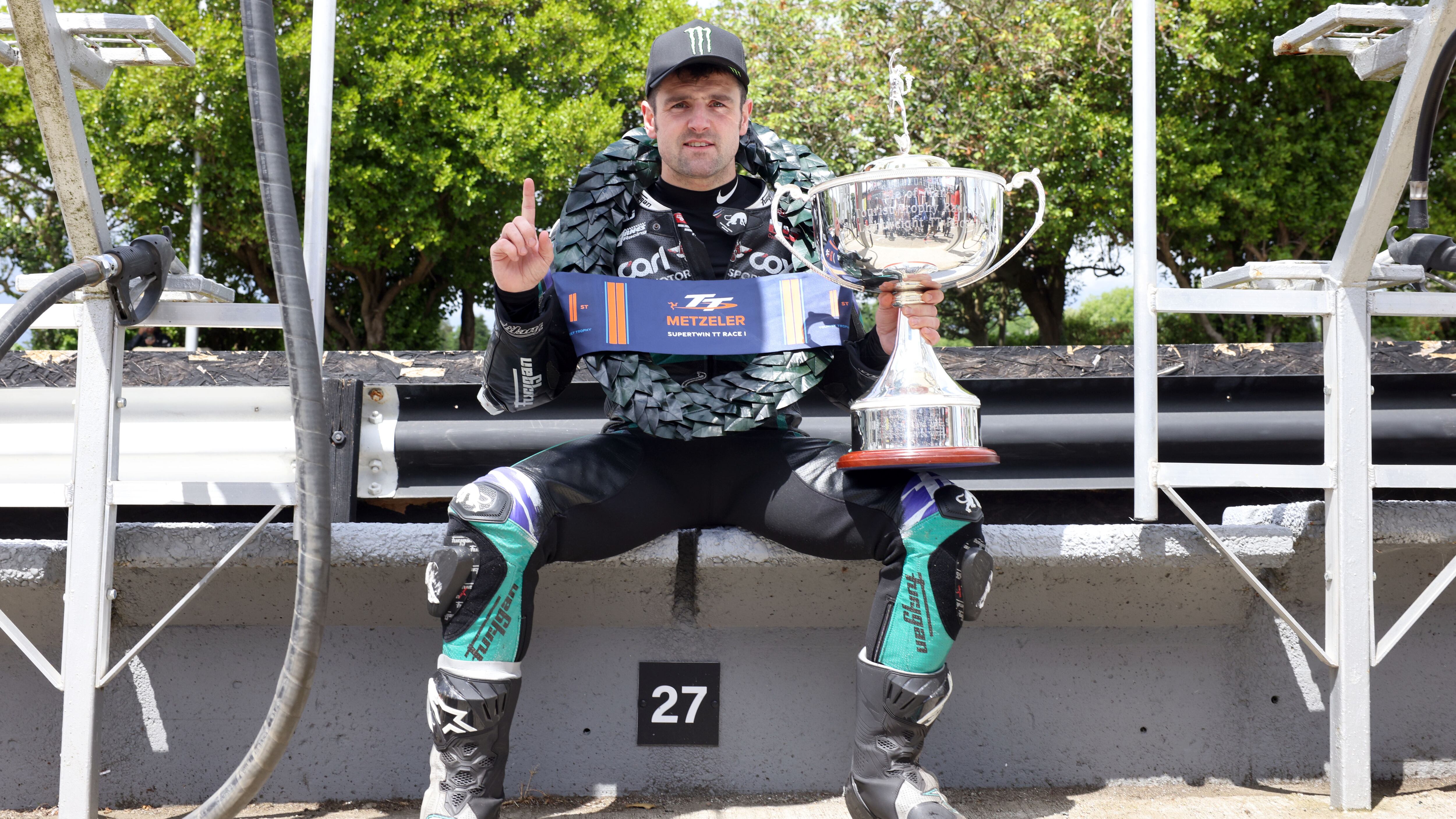 Michael Dunlop sitting with a laurel wreath around his neck and the Metzeler Supertwin trophy in his hand, under him is a sign that says 27
