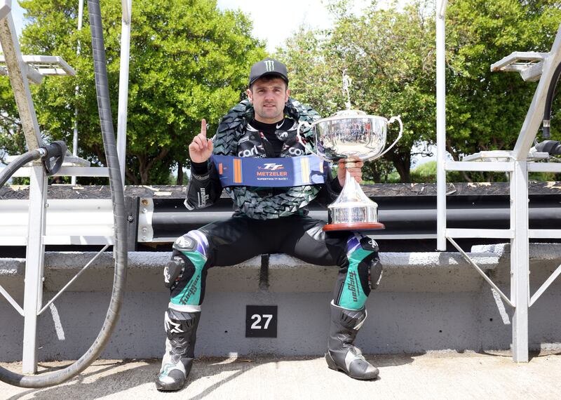 Michael Dunlop sitting with a laurel wreath around his neck and the Metzeler Supertwin trophy in his hand, under him is a sign that says 27