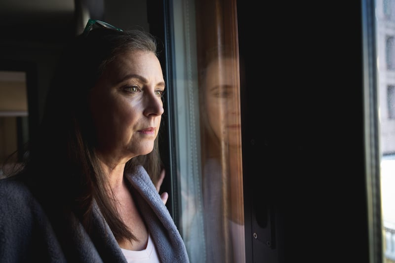 Middle-aged woman with long dark hair staring out of a window looking sad