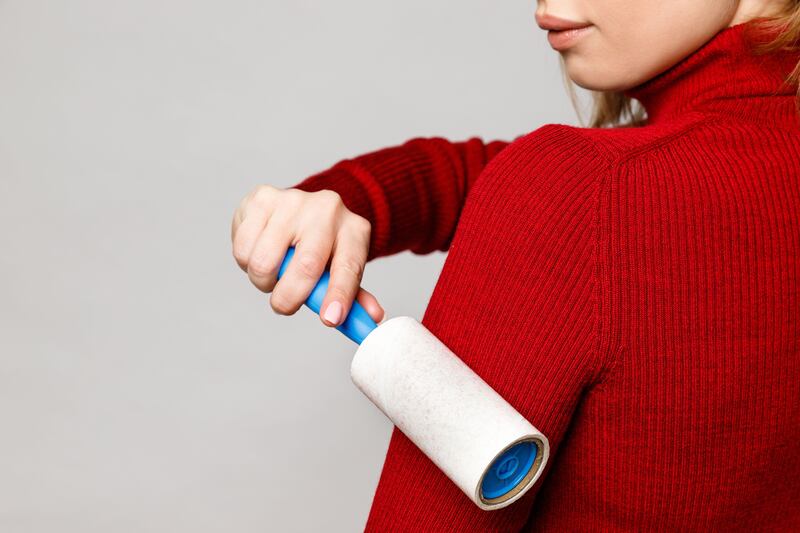 2BF1FRH Woman hand using a sticky roller to clean fabrics – red woolen turtleneck from dust, hair, lint and fluff, front view, close up. Grey background.