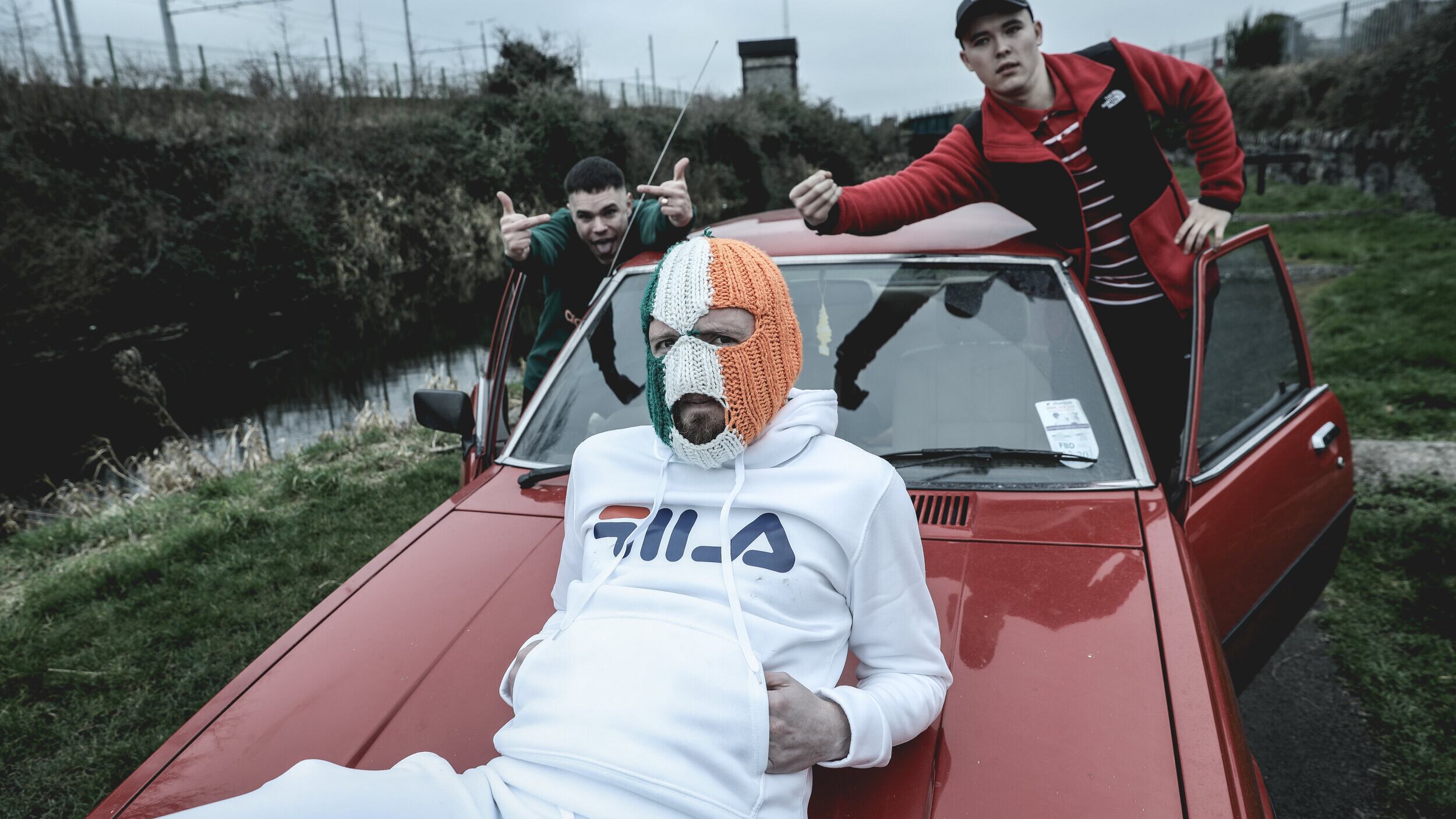 West Belfast rap trio Kneecap pose with a red 1980 Mazda 626