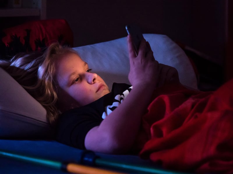 Teenage boy lying in bed staring at his phone in the dark  before falling asleep. Smartphone display is illuminating boy’s face. Caucasian ethnicity.