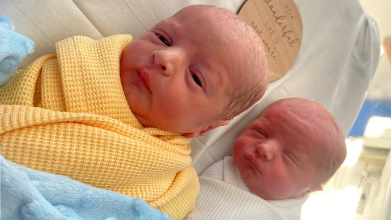 Two babies lying in a hospital cradle, one in yellow swaddling clothes and one in white