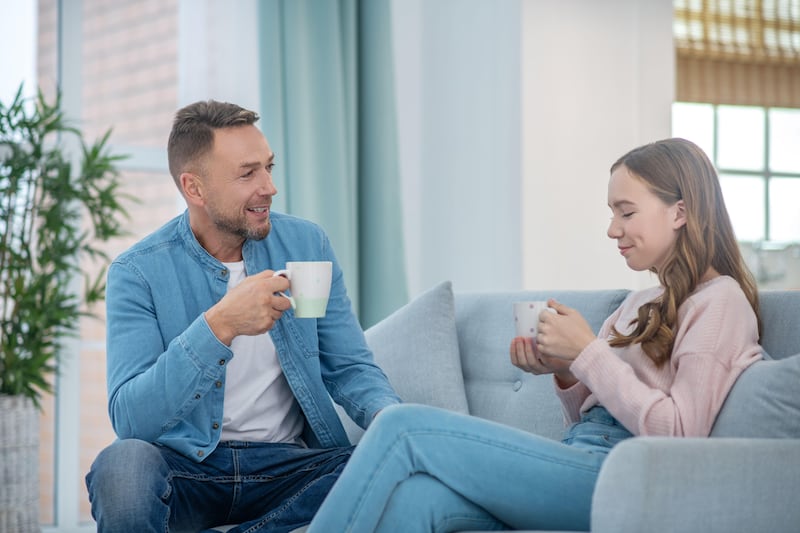 Dad and teenage daughter sitting and chatting on a sofa while holding cups of tea