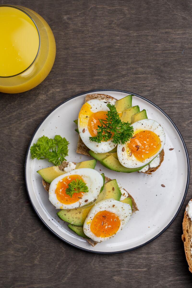 Avocado and boiled eggs on grain bread on a white plate next to a glass of orange juice