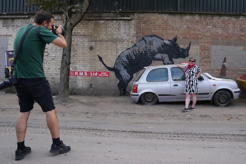 A woman poses for a photograph by a new artwork unveiled by Banksy, depicting a rhinoceros which looks as though it is climbing on top of a car