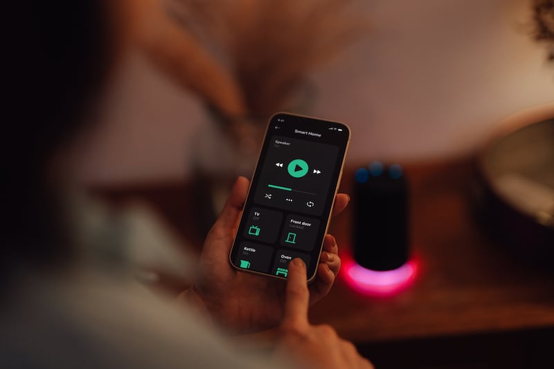 Over the shoulder view of young Asian woman using  smartphone to play music on smart speaker of a modern smart home. Smart home technology concept. Smart living. Lifestyle and technology.