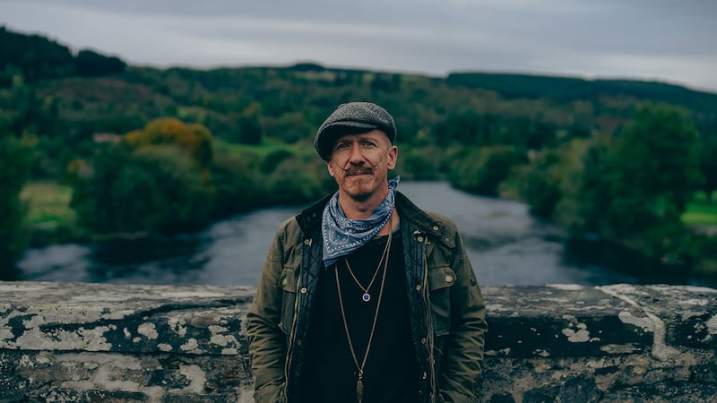 A photo of a moustachioed man standing on a bridge wearing a Barbour jacket, neckerchief and flat cap