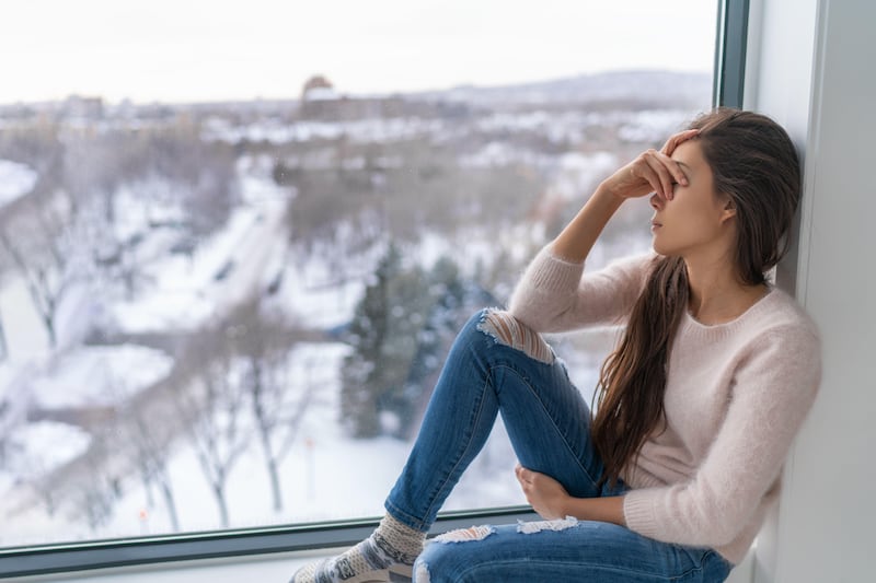 Depressed woman sat on window sill looking out of window at wintry garden