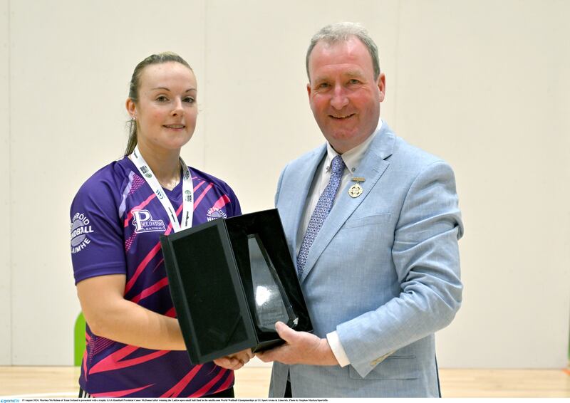 A woman in a purple and pink jersey with a medal around her neck holding a case with a trophy in it and shaking hands with a man in a light grey suit