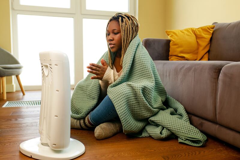 Young woman trying to stay warm by wearing a blanket and sitting next to an electric heater