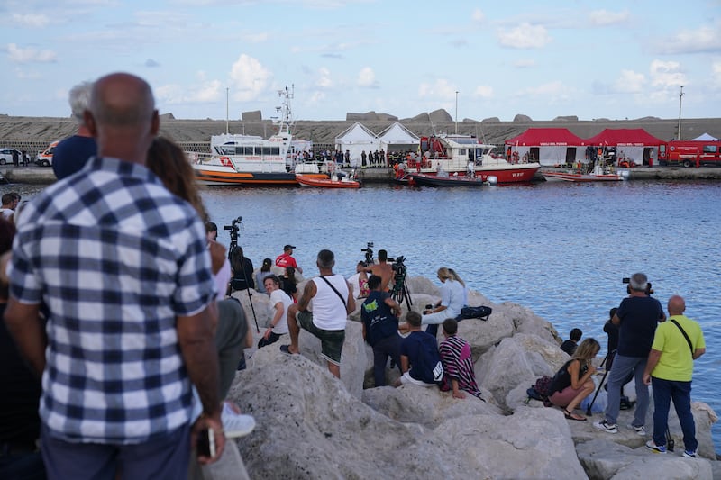 Members of the public and media watch as a body bag is brought ashore at the harbour in Porticello by rescue workers