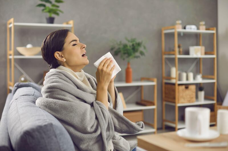 Ill woman wrapped up in blanket sat on a sofa sneezing into a tissue