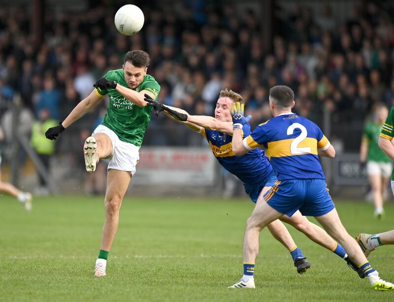 Paul Donaghy wearing a Dungannon kit shooting a gaelic football with a crowd behind him and two gaelic footballers in blue and yellow kits blocking him