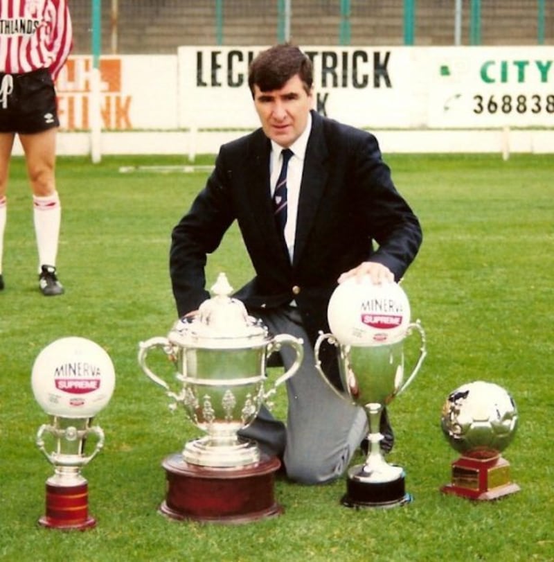 A man wearing a black jacket and tie kneeling in front of four trophies n a pitch with footballs sitting in two of the trophies