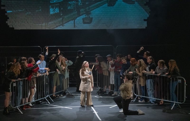 Woman standing in the middle of a stage with a fake microphone- pretending to report the news. There are crowds on her left and right behind a barrier.