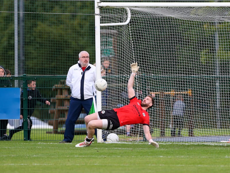 A goalkeeper wearing a red top making a save in a gaelic football match with an umpire looking on