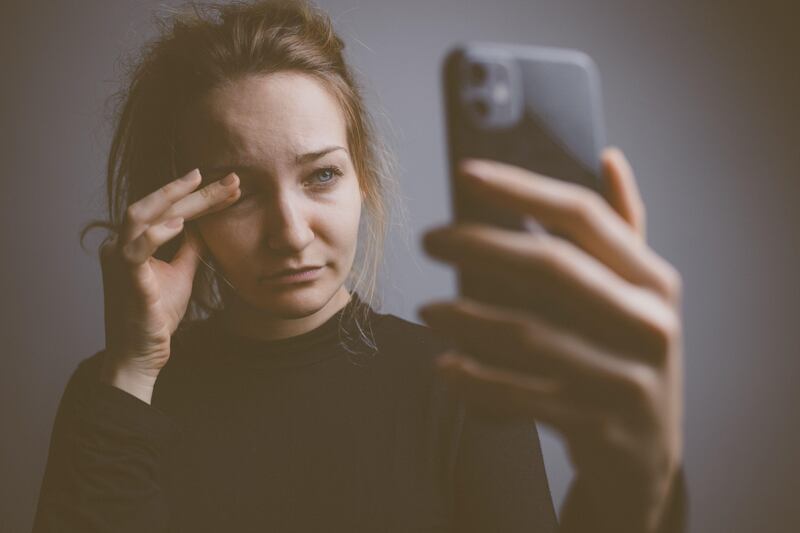 2E55K3Y Young woman holding a phone, tired and exhausted, blue light straining her eyes, messing up her circadian rhytm