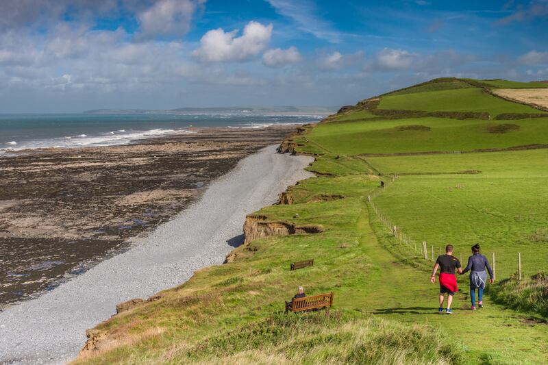 A couple walk along the South West Coast Path, looking towards Westward Ho!, as the sunshine lights up the footpath