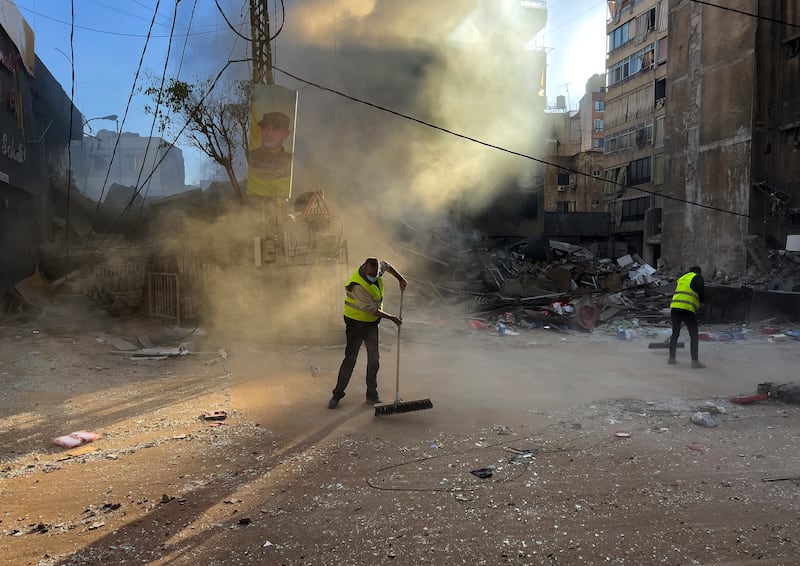 Workers clean a street as smoke rises from a destroyed building that was hit by an Israeli air strike in the southern Beirut suburb of Dahiyeh (Hussein Malla/AP)