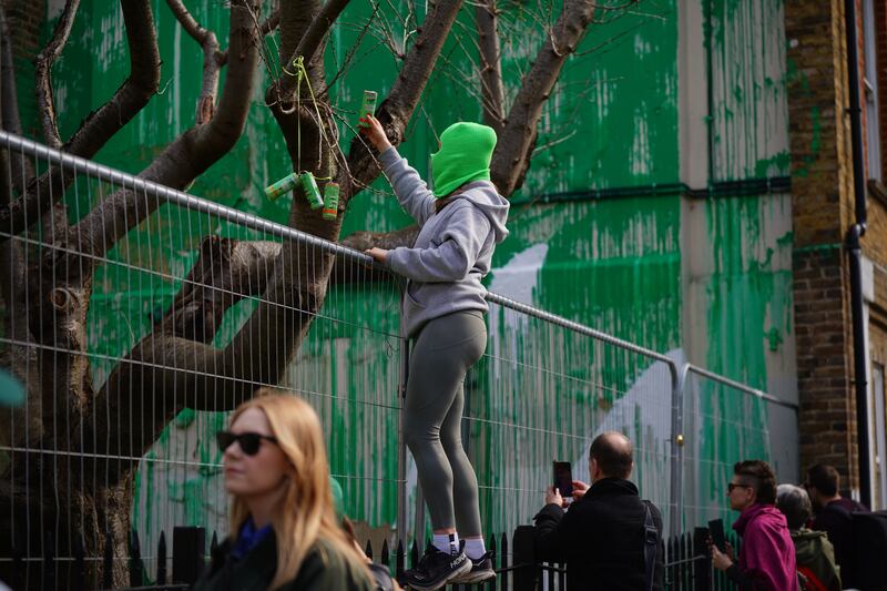 A person ties green cans to the tree in front of the Banksy artwork in Finsbury Park, London after it was defaced with white paint