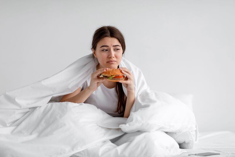 Sad depressed woman sat in bed wrapped in a duvet eating a hamburger