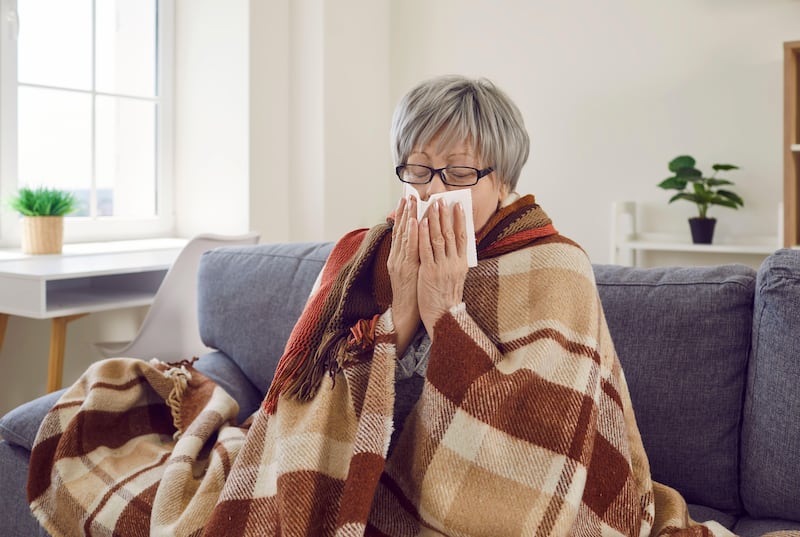 Senior woman with runny nose wrapped in a blanket sat on a sofa