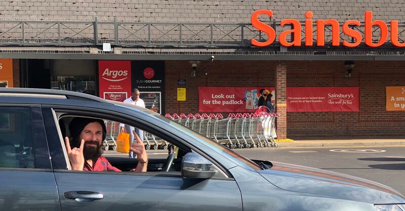 A man sits in a silver car outside a shop which says 'Sainsbury's' and makes the rock sign with both hands