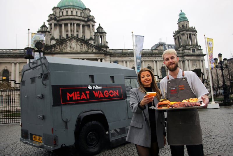 A man and woman holding a tray of food and drink in front of an old RUC land rover with the Meat Wagon Signage parked in front of Belfast City Hall.