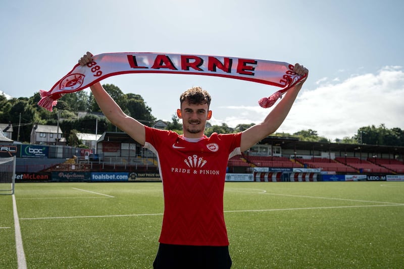 A man standing on a football pitch wearing a Larne FC red football jersey holding up a white scarf that says LARNE on it in red capitals