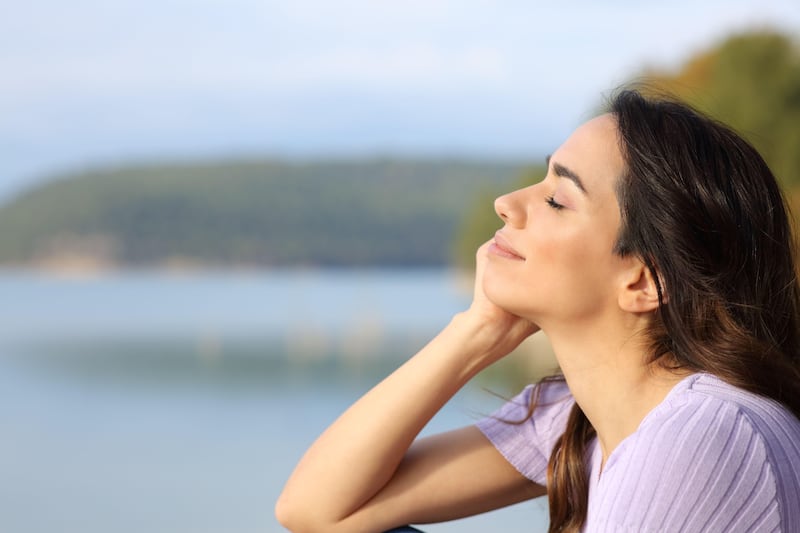 A happy woman closing her eyes and smiling at the sun near a lake