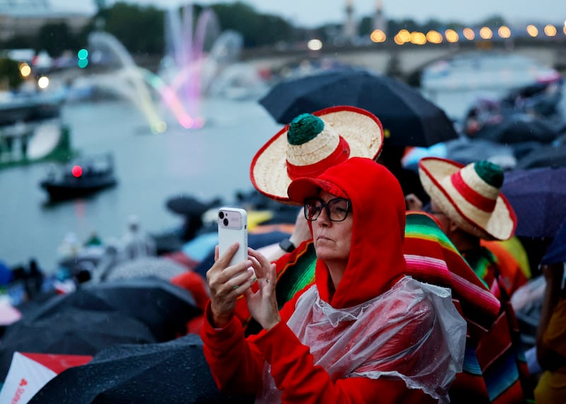 A spectator takes a photo on their phone as athletes ride in a boat along the Seine River during the opening ceremony for the Olympics in Paris (Stefan Wermuth/AP)