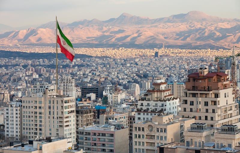 Waving Iran flag above skyline of Tehran at sunset.