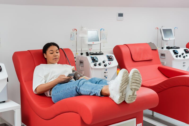 Woman looking at her phone while sitting on a medical chair near transfusion machine while donating blood